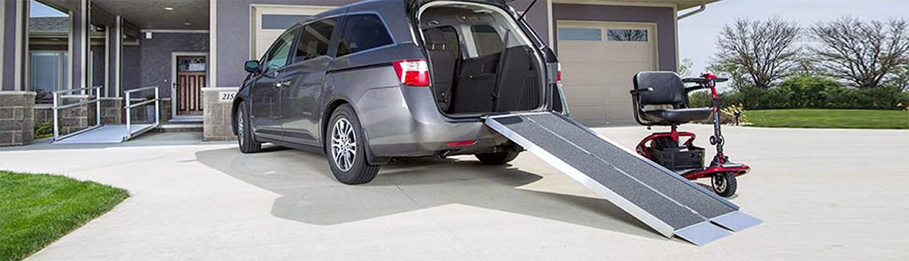 A mobility scooter is parked next to a set of ramps leading into the back of a large car. The weather is sunny.