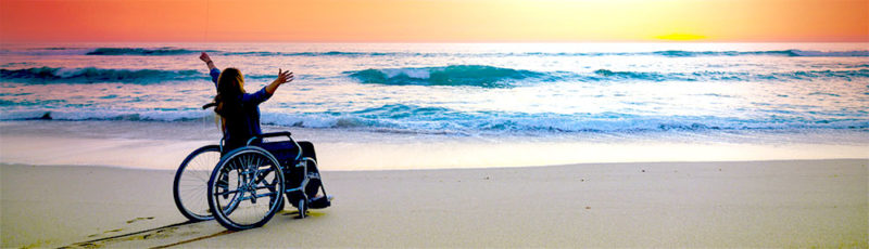 A woman sits on the beach in a wheelchair gazing out to sea admiring the sunset. Her arms are spread wide.